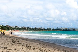 Praia de João Pessoa com guarda-sóis coloridos, ondas e edifícios da cidade ao fundo.