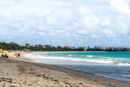 Praia de João Pessoa com guarda-sóis coloridos, ondas e edifícios da cidade ao fundo.