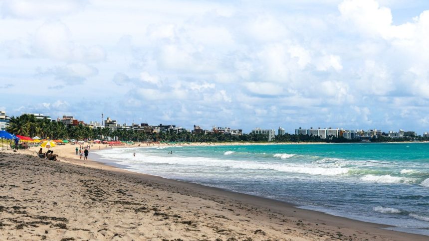 Praia de João Pessoa com guarda-sóis coloridos, ondas e edifícios da cidade ao fundo.