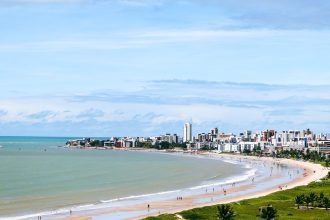 Vista aérea da orla de João Pessoa, com a faixa de areia, o mar e a área urbana ao fundo.