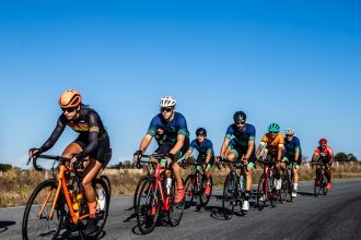 Grupo de ciclistas pedalando juntos ao ar livre sob um céu azul.